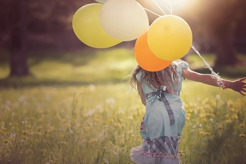 Ein Mädchen von hinten mit mehreren Luftballons am Handgelenk, auf einer Sommerwiese stehend