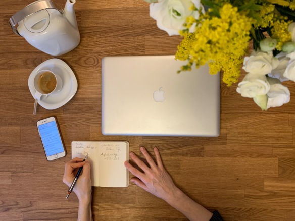 Blick auf Holztisch, auf dem ein Notizbuch, eine Kanne, Espresso, ein Laptop und ein Blumenstrauß zu sehen sind